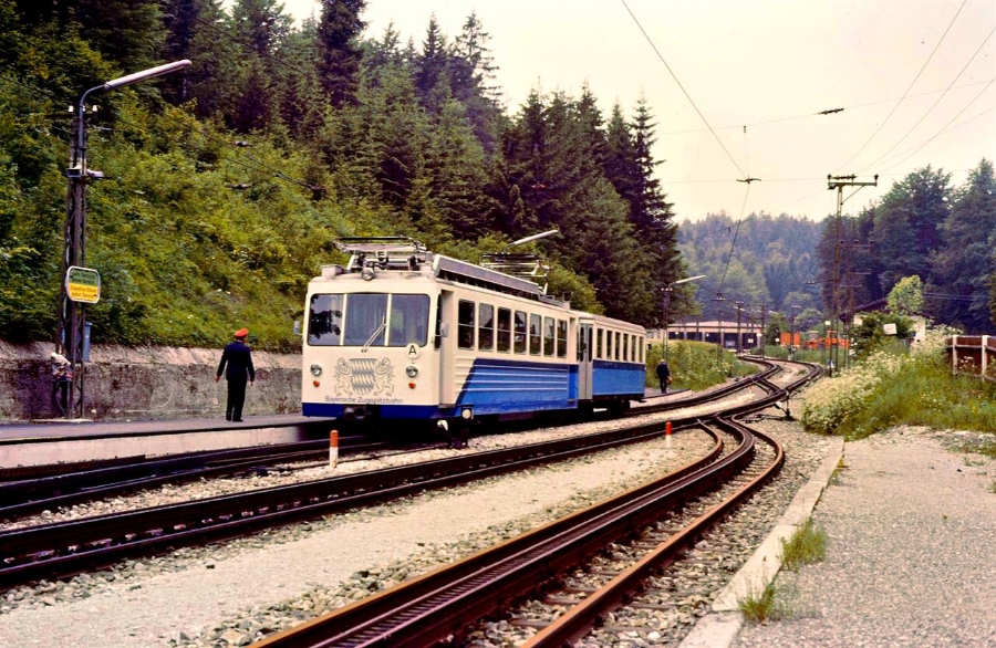 Zugspitze 17 The Bayerische Zugspitzbahn train, painted blue and white, runs along tracks adjacent to a dense forest in Grainau.