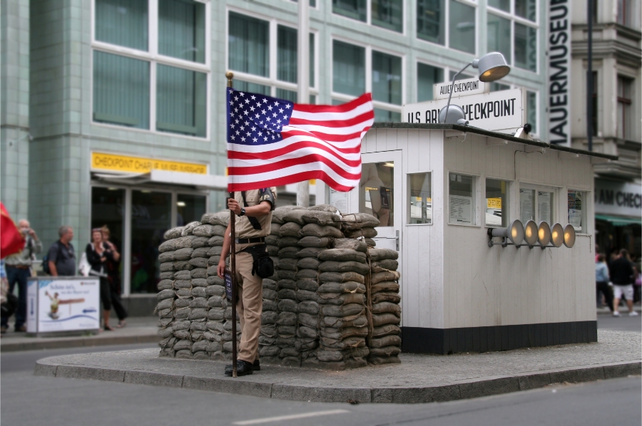 Soldier in uniform stands at Checkpoint Charlie, Berlin, with an American flag visible on the building behind him.