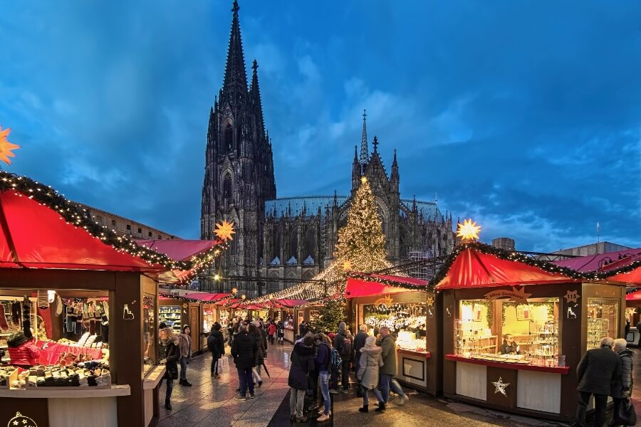 Cologne Cathedral illuminated at night, surrounded by festive stalls at the Christmas market, with visitors enjoying the atmosphere.