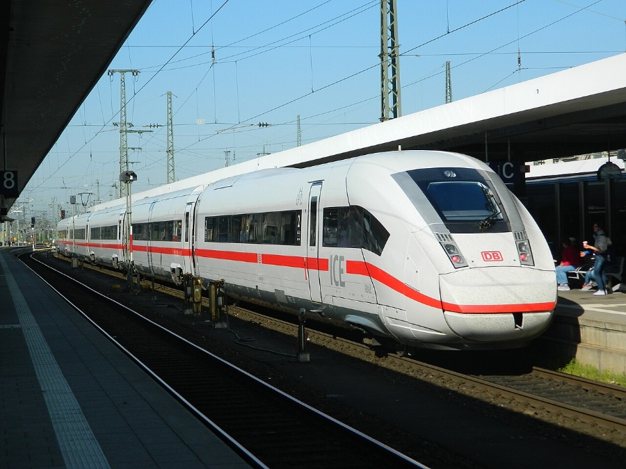 Nuremberg Airport 13 Train arriving at a station, with passengers waiting on the platform and buildings in the background.