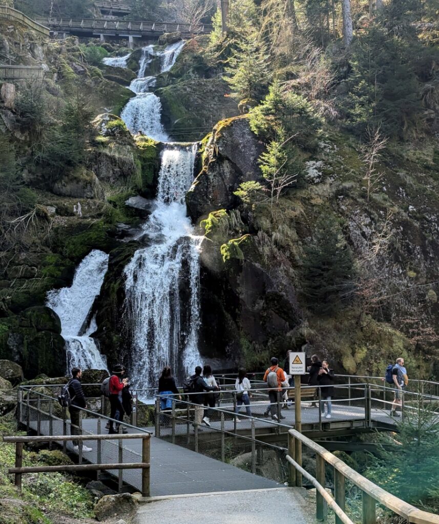 Triberg Waterfalls 6 Tourists stand on a wooden walkway, admiring the cascading Triberg Waterfalls in Germany's lush forest setting.